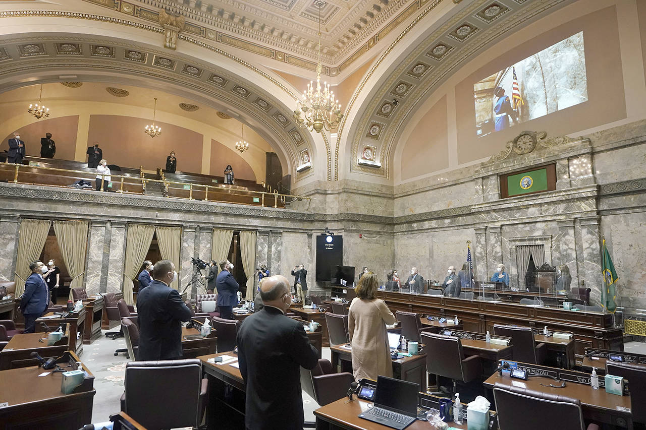 Senate members stand as a ceremonial presentation of colors is done virtually on a video screen above on Monday at the Capitol in Olympia. Washington state’s Legislature convened Monday under a large security presence because of concerns about efforts by armed groups who might try to disrupt the proceedings or occupy the Capitol, which is closed to the public due to the ongoing pandemic. (AP Photo/Ted S. Warren)