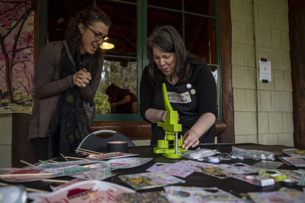 Mayna McVay, 44, left, and “arts and crafts empress” Chris Naqvi, 51, right, complete the final stages of making a button during the Cascadia Tea spring festival at Floral Hall in Everett, Washington on Saturday, April 15, 2023. (Annie Barker / The Herald)
